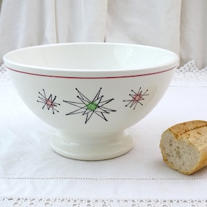 Puede incluir: Tazón de cerámica blanca con un borde rojo y un diseño de estallido de estrellas negro, verde y rosa. El tazón está sobre un mantel blanco con un borde de encaje. Una hogaza de pan está en primer plano.