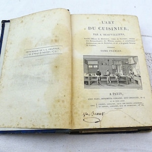 May include: A vintage book titled "L'Art du Cuisinier" by A. Beauvilliers. The book is bound in blue and features a black and white illustration of a kitchen scene on the cover.