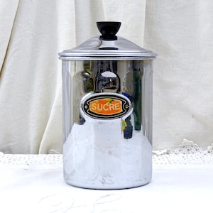 May include: A silver sugar dispenser with a black knob on the lid. The dispenser has a label that says "SUCRE" in orange letters.
