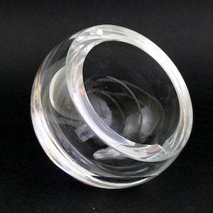 May include: A clear glass bowl with a spherical shape and a thick, curved rim. The bowl features a circular opening and a swirling interior pattern. The transparent glass allows light to pass through. It rests on a black surface.