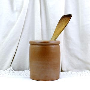 以下が含まれることがあります： 木のスプーンが入った茶色の炻器の壺。円筒形の壺には丸みを帯びた縁があります。スプーンは明るい茶色で、先端は濃い茶色です。背景は白いカーテンです。壺の高さは15〜20cm程度です。