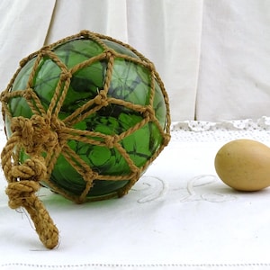 Könnte beinhalten: Ein grüner Glas-Fischerschwimmer, umhüllt von einem geflochtenen Seilnetz, mit einer geknoteten Seilschlaufe. Daneben liegt ein einzelnes, hellbraunes Ei. Die Gegenstände befinden sich auf einer weißen Oberfläche.