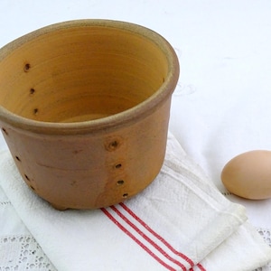 Könnte beinhalten: Ein rustikaler, unglasierter Terrakotta-Käseform mit Abzugslöchern, die auf einem weiß-rot gestreiften Leinentuch liegt. Ein einzelnes braunes Ei liegt neben der Form. Die Form hat eine zylindrische Form.