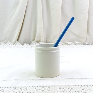Pode incluir: Um pote de cerâmica branco com um lápis azul dentro. O pote é cilíndrico com uma borda ligeiramente alargada. O lápis é azul vibrante e inclinado dentro do pote. O fundo é uma toalha de mesa branca e uma cortina branca.