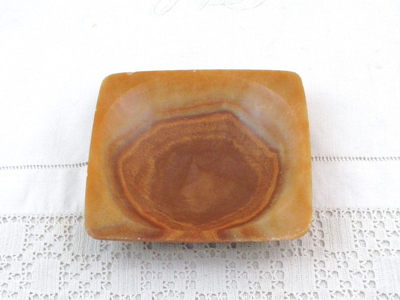 May include: A square, shallow dish made of brown and orange alabaster with a swirling pattern. The dish is empty and has a smooth, polished surface.