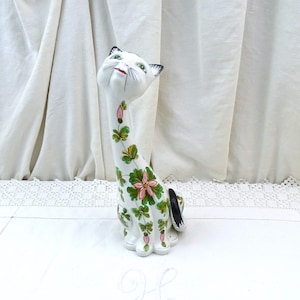 May include: A white ceramic cat figurine with a long neck and a floral design in shades of green and pink. The cat is looking up with a playful expression.