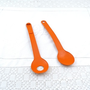 May include: Two orange plastic kitchen utensils. One is a spoon, and the other has a circular hole and a serrated edge. Both have a hole at the end of the handle for hanging. The utensils are on a white surface.