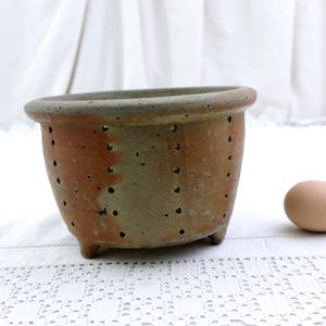 Puede incluir: Una maceta de cerámica hecha a mano, con una estética rústica y terrosa. La maceta es de color marrón apagado con una superficie moteada y texturizada y pequeños agujeros de drenaje. Tiene tres patas pequeñas y un borde redondeado. Hay un huevo al lado.