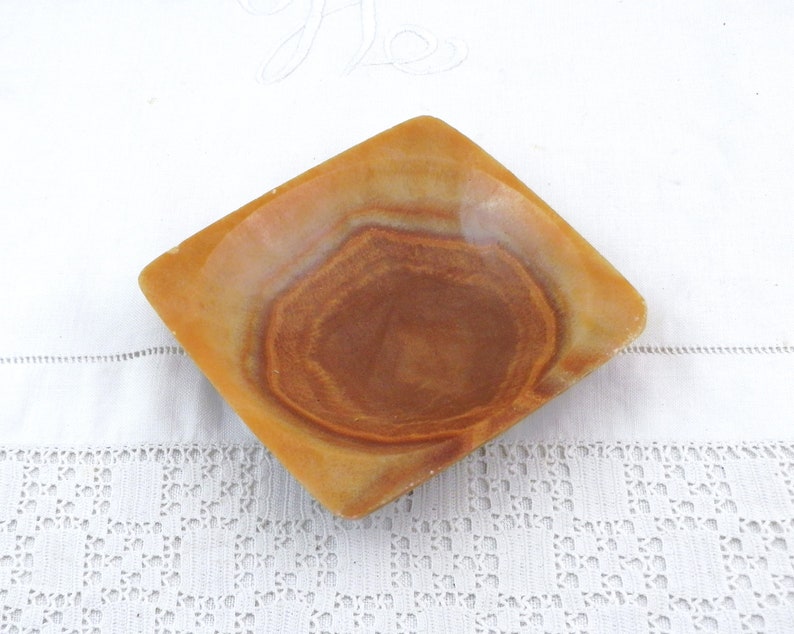 May include: A square, shallow dish made of brown and beige onyx with a swirling pattern. The dish is empty and sits on a white surface.