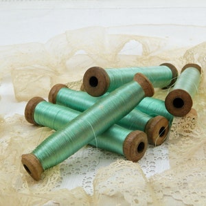 Op de afbeelding: Acht spoelen mintgroen garen, gewikkeld op houten spoelen, liggen op een stuk beige kant.