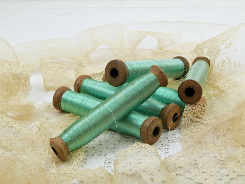 Op de afbeelding: Acht spoelen mintgroen garen, gewikkeld op houten spoelen, liggen op een stuk beige kant.