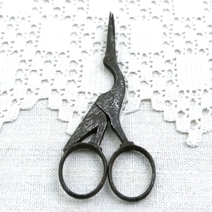 May include: Antique metal scissors with a bird design. The scissors are dark gray with a detailed bird shape, including the head, neck, and body. The scissor handles are circular. The scissors are set against a white, textured background.