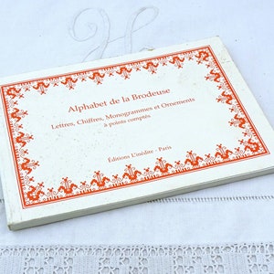 Peut inclure: Un livre blanc avec une bordure orange et le titre "Alphabet de la Brodeuse". Le livre est intitulé "Lettres, Chiffres, Monogrammes et Ornements à points comptés" et "Editions L'inédite - Paris".