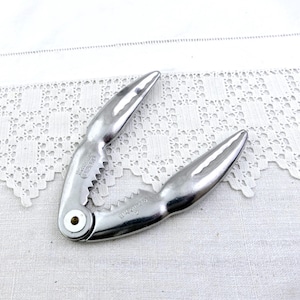 May include: A silver-colored nutcracker with a hinged design. The nutcracker has a serrated edge and pointed tips. The tool is made of metal and is designed to crack nuts. The nutcracker is shown on a white lace surface.