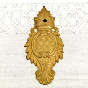 May include: A gold-toned metal decorative accent with a detailed, ornate design. The accent features a central oval shape with a criss-cross pattern and is surrounded by intricate scrollwork and leaf motifs.