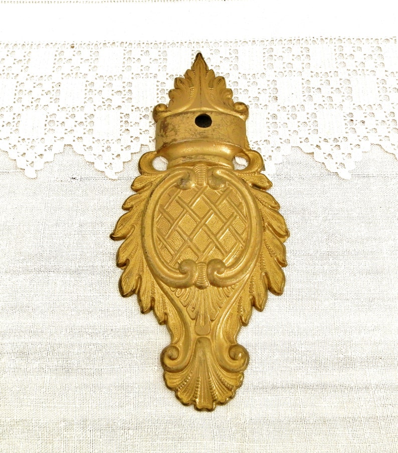 May include: A gold-toned metal decorative accent with a detailed, ornate design. The accent features a central oval shape with a criss-cross pattern and is surrounded by intricate scrollwork and leaf motifs.
