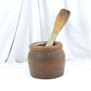 Puede incluir: Un pequeño tarro redondo de cerámica color terracota con una cuchara de madera en su interior. El tarro tiene un aspecto ligeramente envejecido con bandas más oscuras en el centro. La cuchara es de madera clara, mostrando signos de desgaste.
