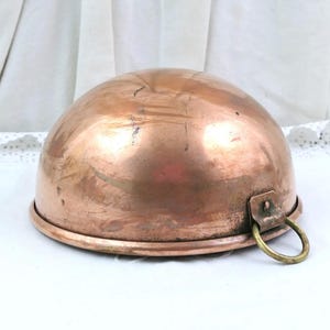 Pode incluir: Uma tigela de mistura semiesférica de cobre com um acabamento metálico polido e sinais visíveis de desgaste. A tigela possui uma única pega em anel de latão fixada na lateral com um pequeno suporte metálico para uma aderência funcional.