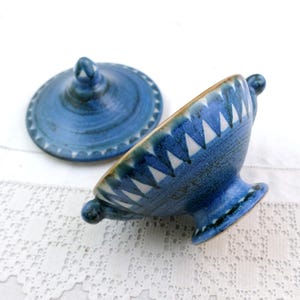 May include: A blue ceramic bowl with a lid, featuring a pedestal base and two small handles. The bowl and lid are decorated with white triangular patterns and a textured finish. The lid is off to the side.