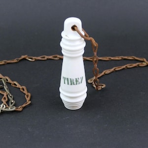 Op de afbeelding: Een witte keramische trekknop voor verlichting met een taps toelopend, geribbeld ontwerp en het woord TIREZ in het groen op de voorzijde gedrukt. De knop is bevestigd aan een lange, roestbruine metalen ketting die rond de basis loopt.