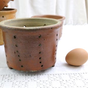 Peut inclure: Plusieurs pots en céramique bruns avec des trous de drainage, parfaits pour la plantation. Un pot est mis au point, avec un œuf brun clair à côté. Les pots ont une surface texturée et sont posés sur un fond blanc.