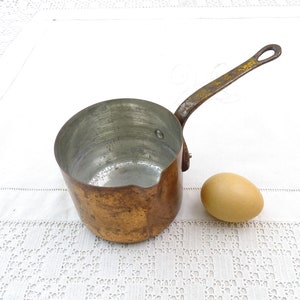 Puede incluir: Una pequeña cacerola de cobre con un mango largo. La cacerola está vacía y tiene un aspecto desgastado y envejecido. Está sentada sobre una superficie de crochet blanco junto a un huevo marrón.