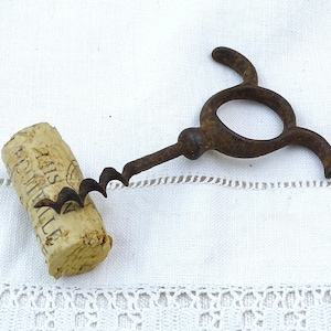 Può includere: Un cavatappi in metallo arrugginito con un designo unico per l'impugnatura. Il cavatappi è appoggiato su una tovaglia bianca con un bordo di pizzo. Un tappo di bottiglia di vino è appoggiato accanto al cavatappi.