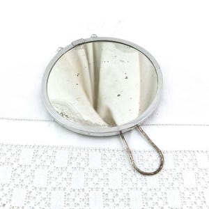 Könnte beinhalten: Ein runder, silberfarbener Handspiegel mit einem glatten Metallrahmen und einem dünnen, geschlungenen Drahtgriff an der Seite. Die reflektierende Glasoberfläche weist Alterserscheinungen mit sichtbaren dunklen Flecken und Verwitterungen auf.