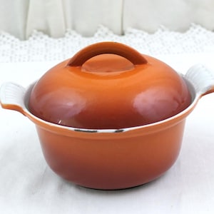 May include: A round, orange enameled cast iron pot with a matching lid and two white, ribbed side handles. The pot features a gradient color finish that transitions from a deep, dark orange at the base to a lighter, vibrant orange at the top.