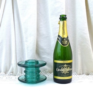 Peut inclure: Un bouchon de bouteille en verre vert avec une base ronde et un dessus cylindrique. Le bouchon de bouteille est posé sur une surface blanche à côté d'une bouteille de champagne Canard-Duchêne Brut.