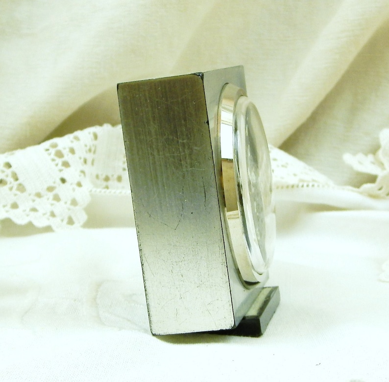 Puede incluir: Un reloj despertador de color plateado con una base negra y una esfera redonda. El reloj est&aacute; sobre una superficie blanca.