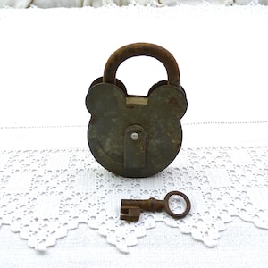 Könnte beinhalten: Ein rostiges Schloss aus Metall mit einem Schlüsselloch und einem passenden Schlüssel. Das Schloss hat eine einzigartige Form mit zwei abgerundeten Ohren an der Oberseite. Das Schloss und der Schlüssel befinden sich auf einer weißen Häkel-Oberfläche.