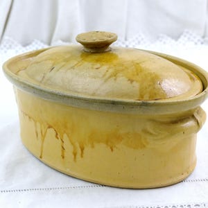 Puede incluir: Cazuela ovalada de cerámica amarilla antigua con tapa. El plato tiene un borde verde claro y un pequeño asa redonda en la tapa. El exterior muestra goteos marrones, lo que aumenta su encanto vintage.