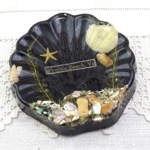 May include: A black, shell-shaped decorative dish with a variety of seashells, a starfish, and the text "Virginia Beach, Va." The dish is filled with small pebbles and other sea elements.