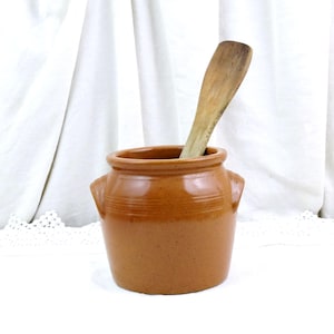 May include: A small, brown ceramic jar with two side handles and a wooden spoon inside. The jar has a rounded shape with horizontal ridges around the middle. The spoon is light brown and appears to be made of wood.