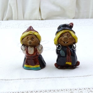 May include: Two ceramic figurines of gnomes, one wearing a red dress and the other wearing a blue coat. Both have yellow hair and are smiling.