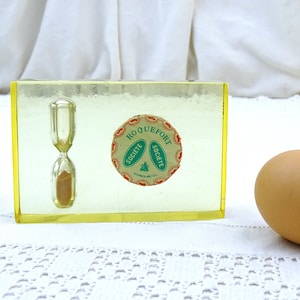 Puede incluir: Un reloj de arena de plástico amarillo con una etiqueta circular que dice "Roquefort Societe". El reloj de arena está sobre una superficie de crochet blanco junto a un huevo marrón.