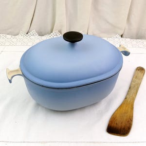 Puede incluir: Una olla ovalada azul claro con un pomo negro y asas color crema. Una espátula de madera descansa junto a la olla. La batería de cocina está sobre una superficie blanca.