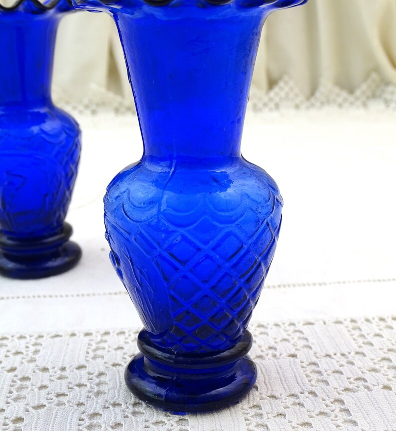 May include: Two cobalt blue glass vases with a diamond pattern and scalloped rims. The vases are on a white crocheted surface.