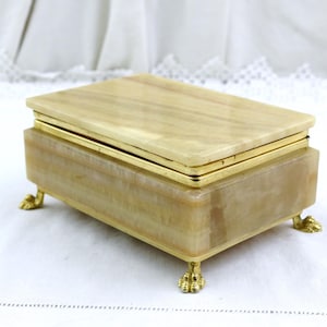 Könnte beinhalten: Eine rechteckige Schmuckschatulle mit einem hellbeige Onyx-Korpus und einem goldfarbenen Metallrahmen. Die Schatulle steht auf vier kleinen, dekorativen Klauenfüßen. Der Deckel ist ebenfalls aus Onyx, mit goldfarbener Metallverkleidung.