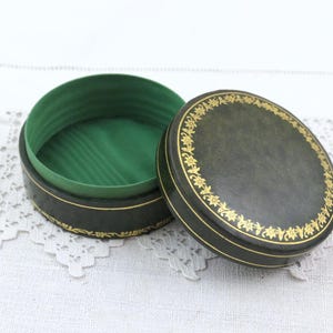 May include: A round, dark green lidded box with a gold floral border pattern on the lid and base. The interior of the box is lined with a bright green fabric, and the lid is placed beside the open base, revealing the green interior lining.