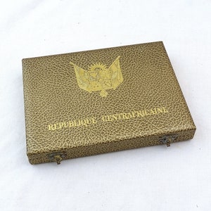 Puede incluir: Una caja de color dorado con una superficie texturizada y las palabras "REPUBLIQUE CENTRAFRICAINE" impresas en la parte delantera. La caja tiene un emblema dorado con un sol y una bandera.