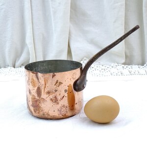 May include: A small copper saucepan with a long, curved handle. The saucepan has a hammered finish and is sitting on a white surface next to a brown egg.
