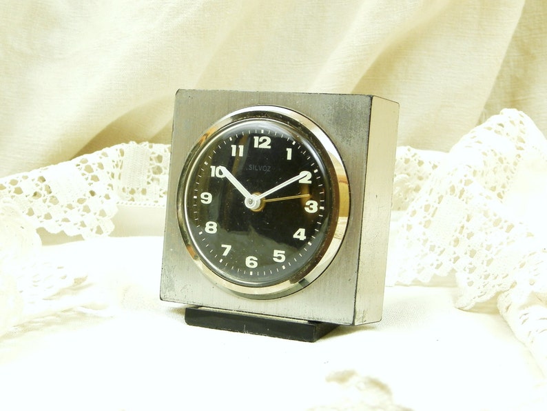 Puede incluir: Un reloj despertador cuadrado plateado y negro con una esfera negra. El reloj tiene una caja de metal plateado y una base de pl&aacute;stico negro. La esfera del reloj tiene n&uacute;meros y manecillas negros, y la palabra "Silvox" est&aacute; impresa en blanco en la esfera.