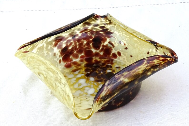 May include: A square-shaped glass bowl with a mottled brown and yellow pattern. The bowl has a unique, wavy design and a slightly flared rim.