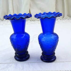 May include: Two cobalt blue glass vases with a ruffled top and a floral design. The vases are on a white tablecloth.