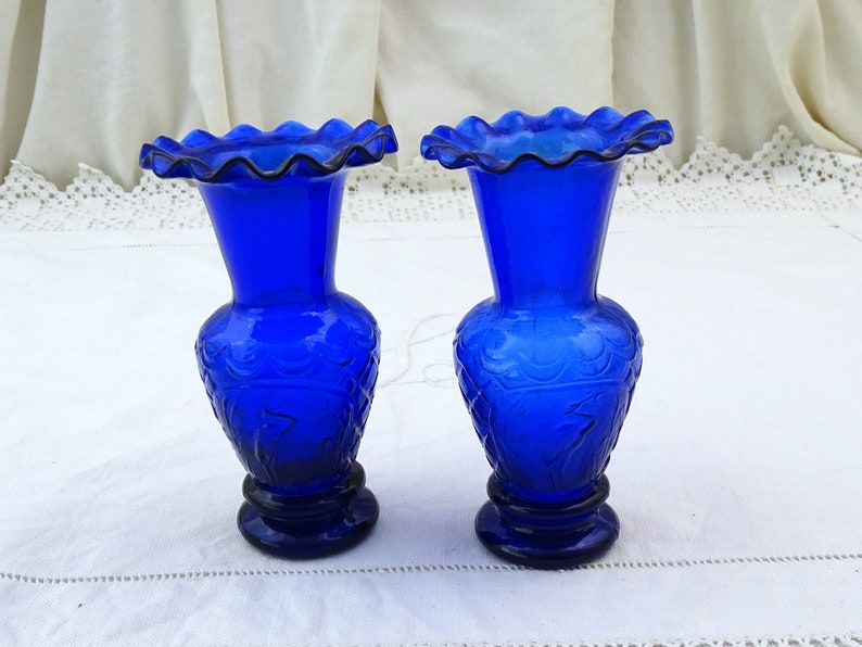 May include: Two cobalt blue glass vases with a ruffled top and a floral design. The vases are on a white tablecloth.