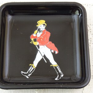 May include: Black square ashtray with a white and red image of a man in a yellow hat and red jacket walking with a cane.