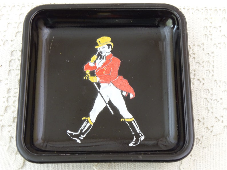 May include: Black square ashtray with a white and red image of a man in a yellow hat and red jacket walking with a cane.
