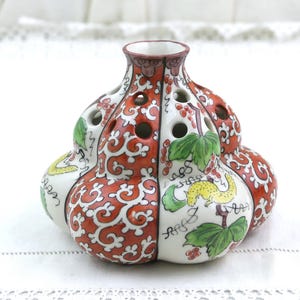 May include: A ceramic flower frog vase with a bulbous, multi-lobed shape. The surface features alternating panels of intricate orange and white scrollwork and white sections painted with green leaves, red berries, and a small yellow creature.